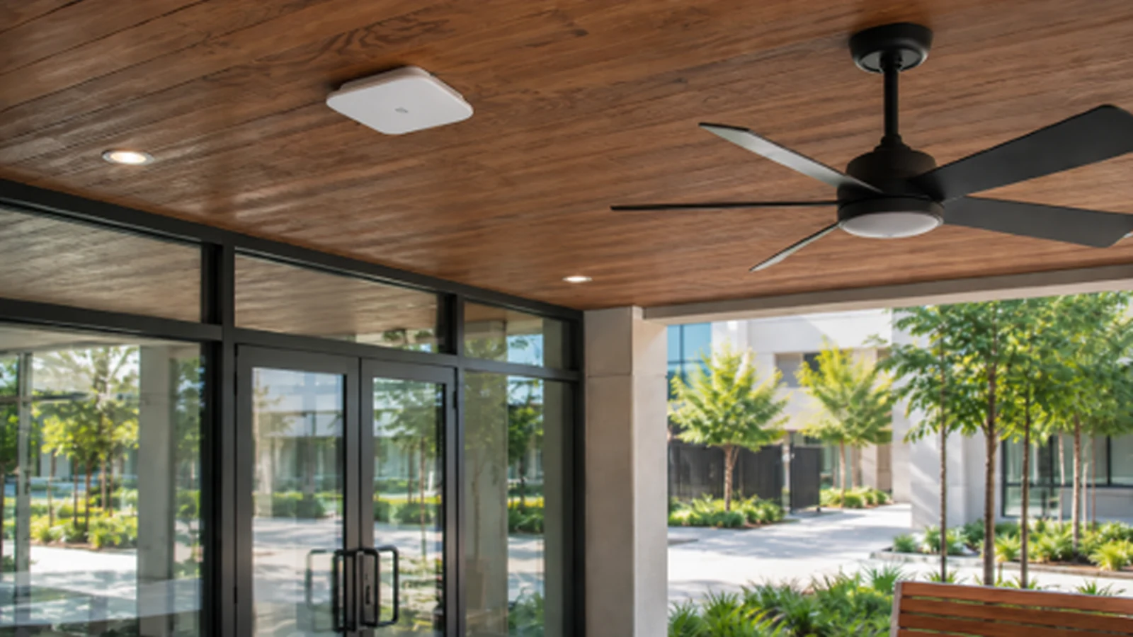 Commercial wireless access point mounted on a wood soffit beside a ceiling fan and glass entrance.