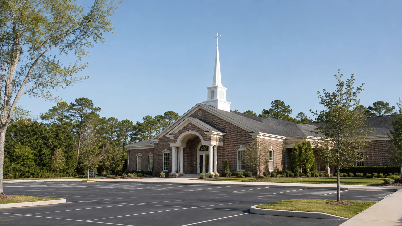 Completed brick church building and steeple viewed across its landscaped parking area in daylight.