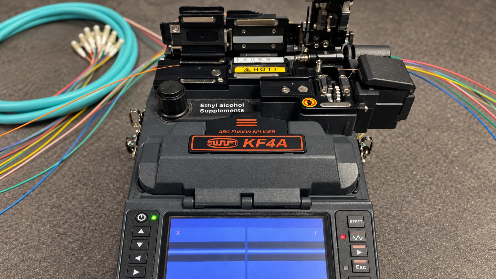 Fusion splicer with open fiber-alignment mechanisms and prepared optical fibers on a workbench.