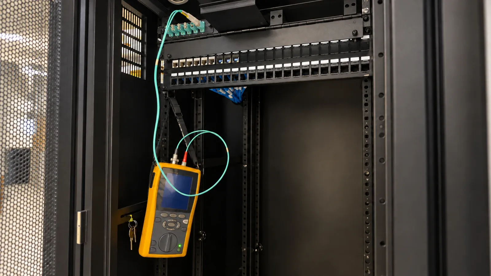Handheld optical tester supported by a strap, with aqua test leads connected to a rack fiber panel.